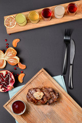 Flat lay of meat steak, pomegranate, tangerine, alcohol cocktail drinks on black background, top view with copy space.