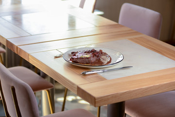 Grilled meat steak served on white plate with utensils on light wooden table in restaurant. Restaurant food photography