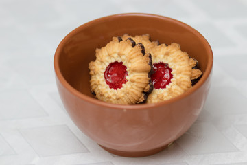 Crispy dough cookies stuffed with strawberry jam, with a chocolate-coated bottom. Inside a red pottery bowl. Against a blurry white background.