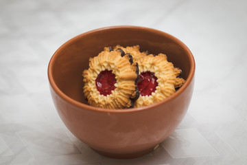 Crispy dough cookies stuffed with strawberry jam, with a chocolate-coated bottom. Inside a red pottery bowl. Against a blurry white background.