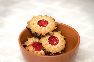 Crispy dough cookies stuffed with strawberry jam, with a chocolate-coated bottom. Inside a red pottery bowl. Against a blurry white background.