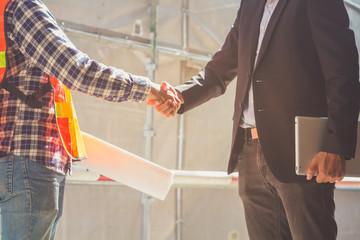 A businessman hold notebook and an engineer hold drawing plan shaking hands, successful in business negotiation deal , finishing up a project meeting at site construction. Handshake Business concept.