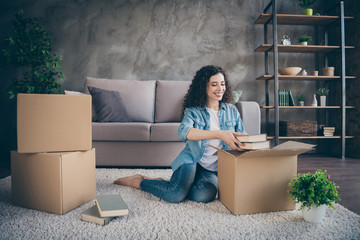 Nice attractive lovely cheerful cheery wavy-haired girl sitting on floor carpet opening unwrapping unpacking her own things in modern loft industrial style interior living-room