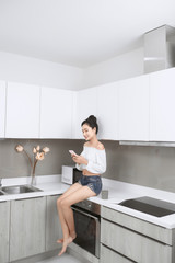 Fototapeta na wymiar Young beautiful Asian woman using smartphone and smiling while sitting in kitchen