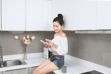 Beautiful Asian woman holding credit card and smartphone while sitting in kitchen.