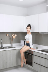 Portrait of Cheerful woman sits on countertop in white modern kitchen.