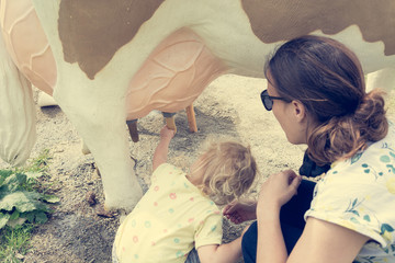 Cute blonde girl learning how to milk a cow on milking simulator with her mother.