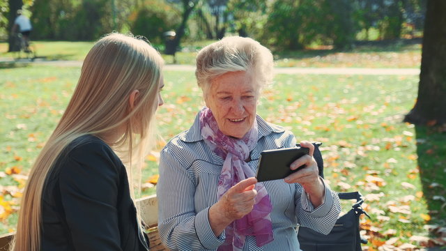 Beautiful blonde girl with elder woman sitting on bench and looking something on smartphone. They spending time in a big autumn park with golden leaves on a grass.