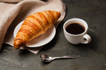 Croissant and cup of coffee with spoon and saucer on a dark background with gray linen cloth