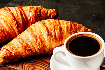 Two delicious, juicy croissants and cup of espresso with foam and spoon on a dark wooden plate