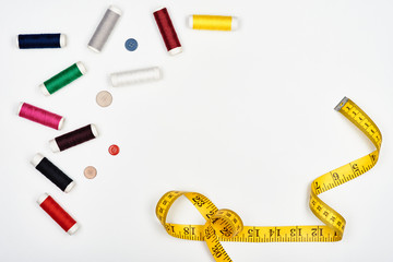 Spools of colorful thread for sewing, multi-colored buttons and tape measure on a white surface. Top view.