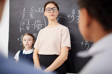 Young teacher in eyeglasses standing near the blackboard with formulas and listening to school children
