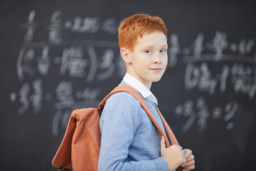 Portrait of red haired schoolboy standing with backpack behind the back and looking at camera with blackboard in the background