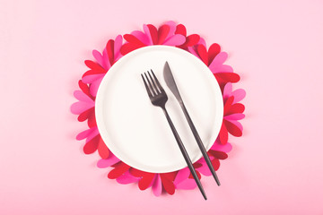 Round white plate with a black fork and a knife on a pink table with paper hearts. Valentine's day concept and laid table.