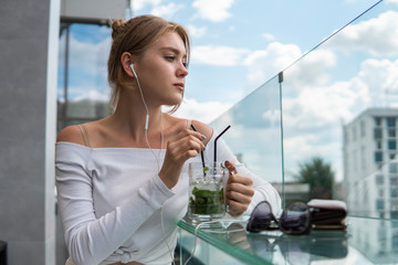 Cheerful blond woman blogger with cocktail is listening music or e-book on smartphone connected to free wifi in cafe. Positive young student listening podcasting online on modern mobile phone.