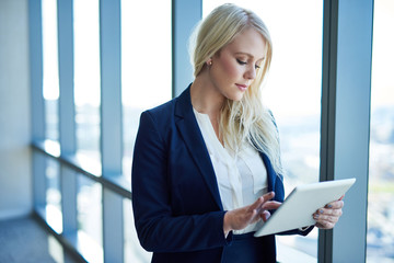 Focused young businesswoman standing by office windows using a tablet