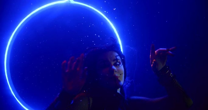 A beautiful femme fatale with unusual makeup and long hair is in a dark Studio under water on a blue background. Her hair is flying. Behind it is a glowing circle.