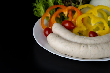 Two long white raw bratwurst sausage with mix vegetables salad in the white plate isolated on black background