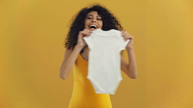 happy bi-racial pregnant woman showing romper isolated on dark yellow