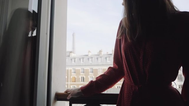 Close-up slide on beautiful woman in red dress standing at French balcony window enjoying Eiffel Tower view slow motion.