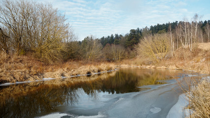 River with ice surrounded by forests and dry grass in snowless winter autumn or spring under a blue sky