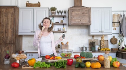 Pregnant Woman Dancing And Singing In Headphones With Mobile Phone On Kitchen.
