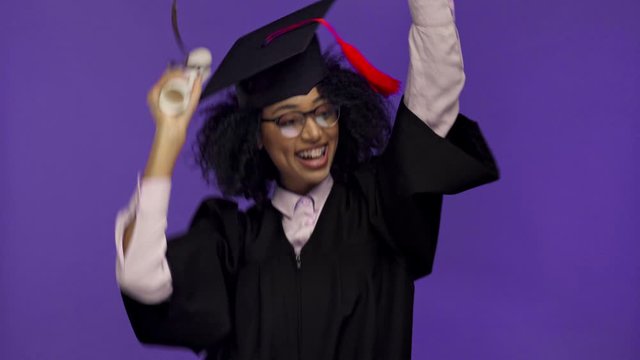 smiling african american graduate dancing with diploma isolated on purple 