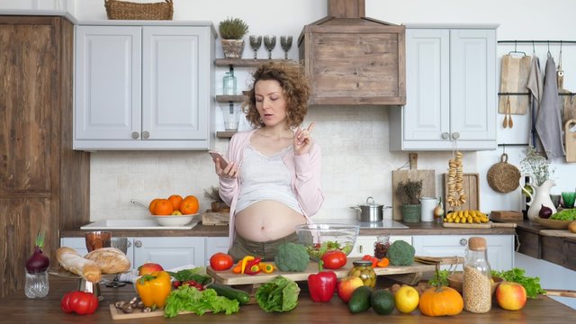 Happy Pregnant Woman Cooking Healthy Food On Kitchen And Using Cell Phone.
