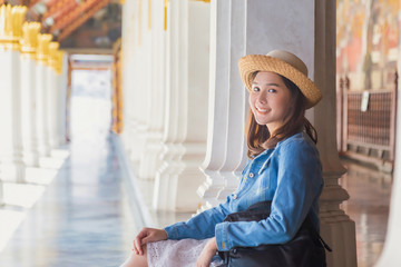 An Asian woman traveler in white dress and jean jacket under straw hat sitting on floor, smile on face and backpack