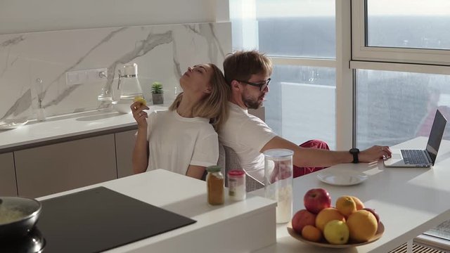 Side view of bearded man working on laptop computer sitting at home on a kitchen with panoramic windows. Young blonde wife sitting from behind and enjoying eating her apple in the morning