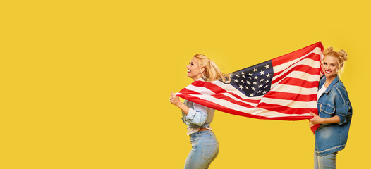 American girls. Happy young women in denim clothes holding USA flag isolated on yellow background....