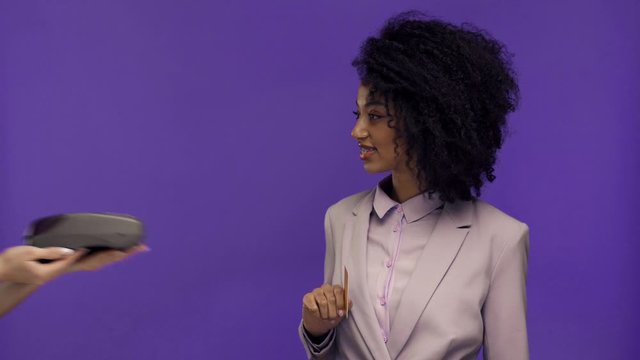 cropped view of woman holding terminal and african american businesswoman paying with credit card isolated on purple 