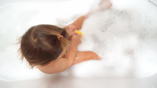 Baby girl washing in bath with foam and yellow rubber duck