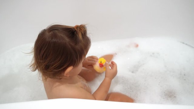 Baby girl washing in bath with foam and yellow rubber duck