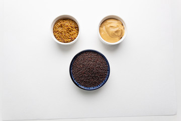 Cream consistency yellow mustard, seeds with vinegar in white bowls and black indian mustard on white background