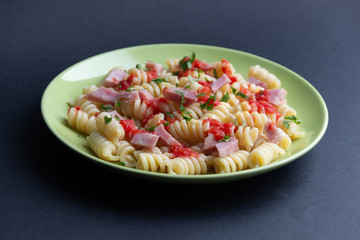 Hot fusilli pasta with tomato, bacon, cheese, greenery on green plate on black background