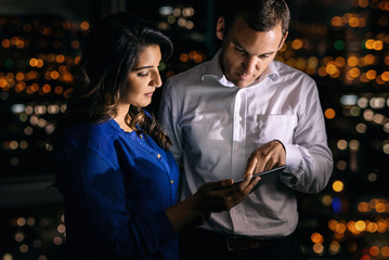 Two coworkers using a digital tablet while standing together in an office late at night in front of windows overlooking the city