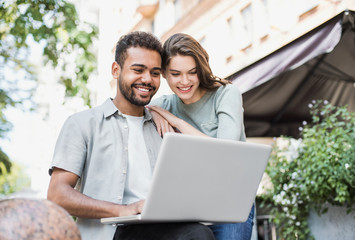 Beautiful happy couple using laptop. Joyful smiling woman and man looking at gadget in a city in summer. Love, relationship, technology and communication concept