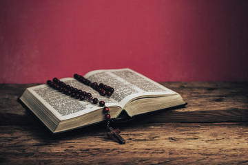Open Holy Bible and beads crucifix on a old oak wooden table. Beautiful red wall background.