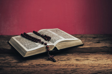 Open Holy Bible and beads crucifix on a old oak wooden table. Beautiful red wall background.