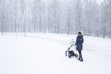 Young mother pushing baby stroller and walking at nature park. Snow covered trees. Spending time with infant in beautiful snowy winter day. Enjoying peaceful stroll. Back view.