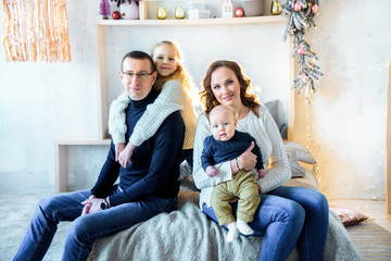 Happy mom and dad, sister and little brother on the bed in a large bright bedroom. Christmas decor and lights. Big happy family. The concept of parental love and parenting