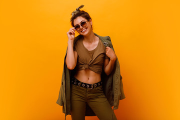 Fototapeta premium Full length Young fashion,stylish smiling brunette with bandana hair roll woman in the studio. Fashion trend apparel, safari style .Girl in high spirits in khaki clothes posing on orange background