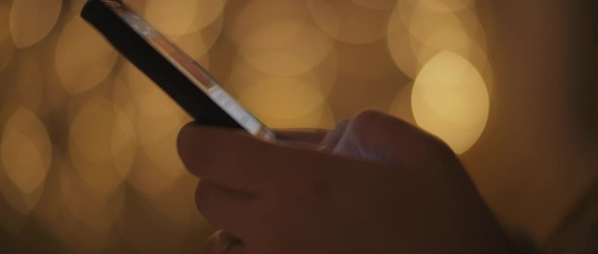 Close up of a young woman's hands typing a message, texting on her phone,  with bokeh lights on the background. Shallow dof, realtime, BMPCC 4K