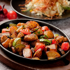 Delicious oven-baked potatoes with herbs and vegetables in a pan, rustic style. Potatoes fried, red cherry tomato, zucchini, onion on gray stone background.