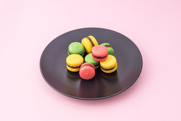 Obraz na płótnie Canvas Tasty french macarons in a black plate on pink pastel background. Red, yellow and green macarons