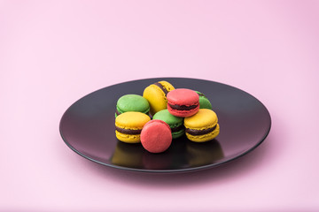 Tasty french macarons in a black plate on pink pastel background. Red, yellow and green macarons