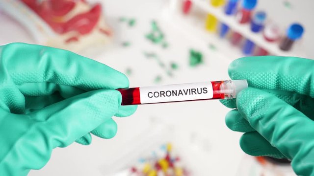 Laboratory scientist showing to the camera a blood test tube containing the Coronavirus. Macro closeup shot of the vial. Wearing gloves and a biohazard hazmat suit.