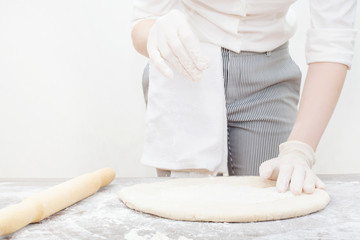 Obraz na płótnie Canvas The cook makes the dough, on the table with sprinkled flour, in working white gloves, in a professional uniform, on a white background. Concept of making pizza.