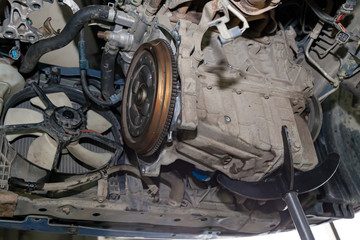 Fototapeta premium A car raised on a lift for repair and under it a detached Transmission gearbox suspended on a crane and a gear box on a lifting in a vehicle repair shop with view to the bottom in vehicle workshop.